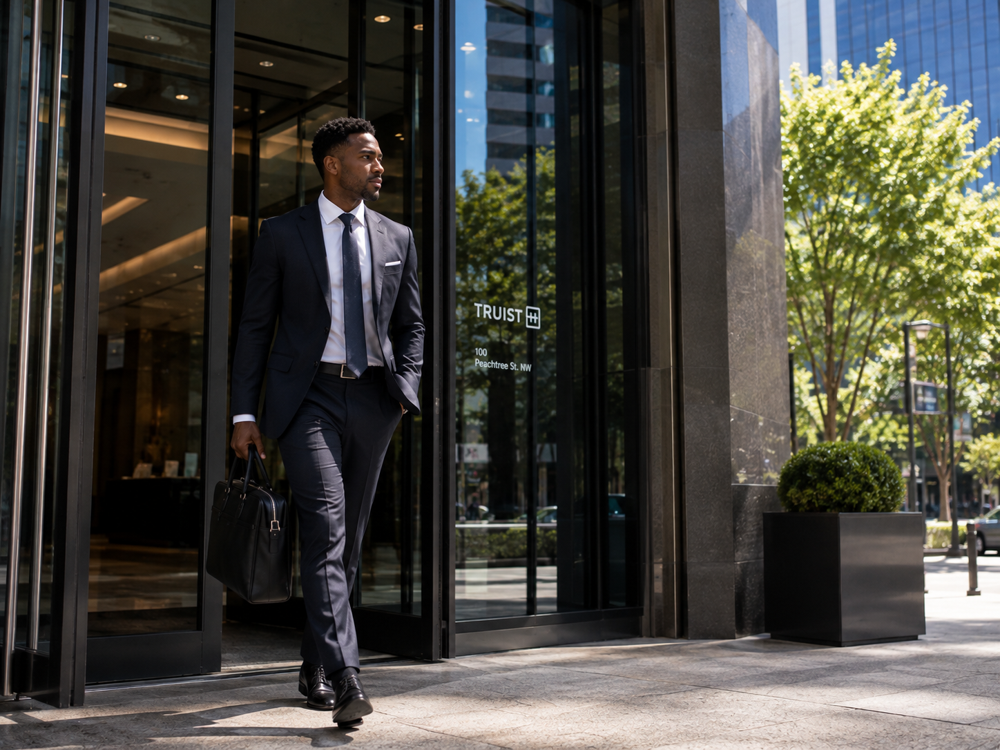 A man in a dark navy suit, white dress shirt, and tie walks out of a modern building entrance carrying a black briefcase. The background shows glass doors, reflections, and green trees.
